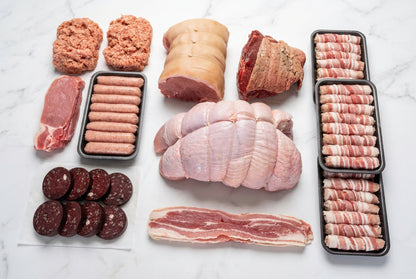 An assortment of christmas meats including sausage meat, bacon, sausages, black pudding, a gamon roasting joint, a beef rump roasting joint, streaky bacon, 3 trays of pigs in blankets, a whole 5kg boneless turkey butterfly strung arranged on a white background.