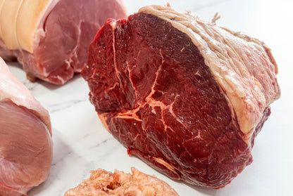 Raw beef rump roast, with a gammon joint in the background arranged on a white surface, and sausage meat in the foreground.