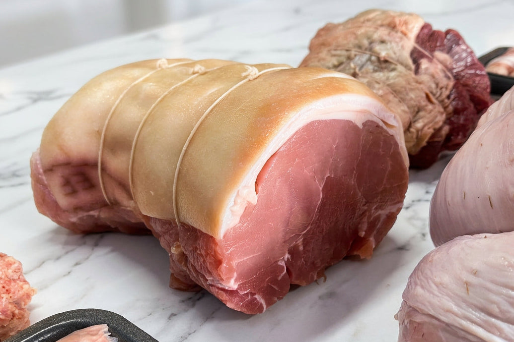 Fresh raw meats on a white table, including a tied gammon roast, a cut of beef rup roast, a strung turkey butterfly in the right foreground, and sausage links in black trays.