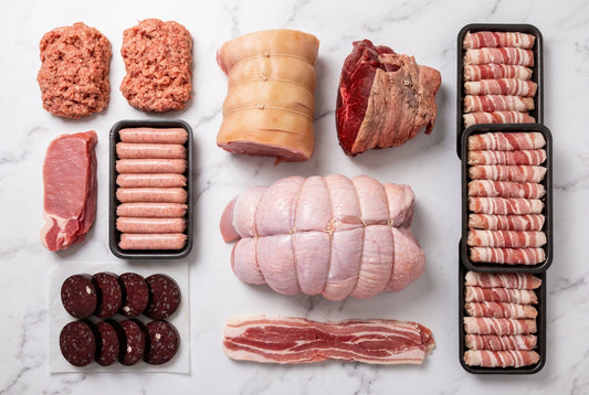 An assortment of christmas meats including sausage meat, bacon, sausages, black pudding, a gamon roasting joint, a beef rump roasting joint, streaky bacon, 3 trays of pigs in blankets, a whole 5kg boneless turkey butterfly strung arranged on a white background.