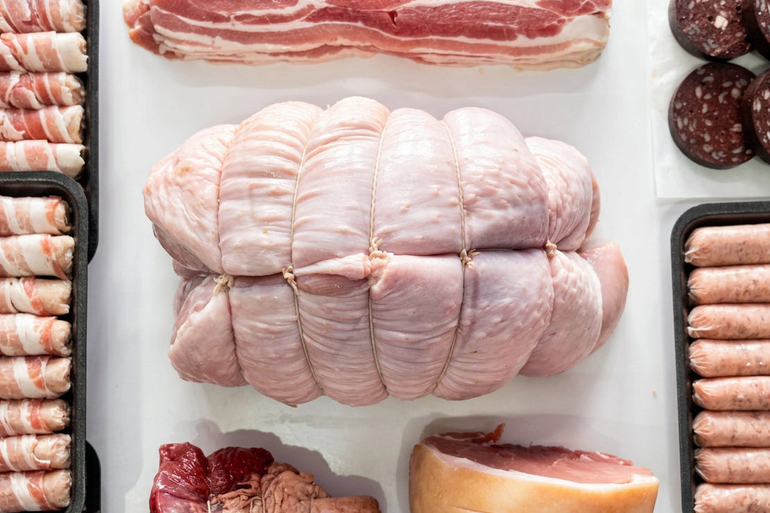 A raw, tied 5kg whole boneless turkey butterfly roast displayed on a white surface, surrounded by other cuts of meat including gammon, rump roast, sausage, and pigs in blankets