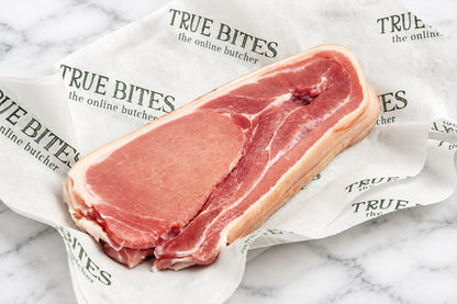 A raw cut of middle bacon resting on branded white paper over a dark wooden surface. The paper is printed repeatedly with the brand name 'TRUE BITES the online butcher'.