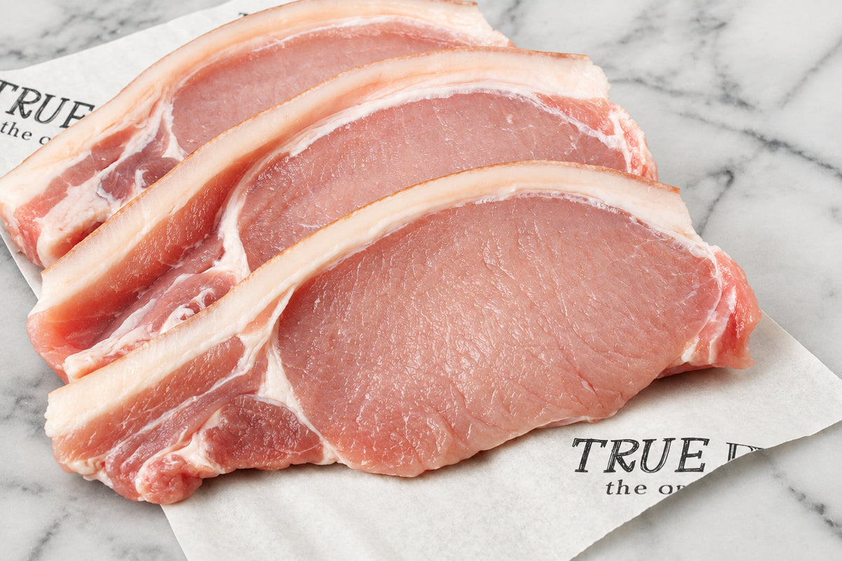 three raw pork chops with rind and visible fat layers, arranged on white butcher paper over a dark wooden surface.