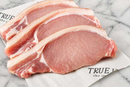 three raw pork chops with rind and visible fat layers, arranged on white butcher paper over a dark wooden surface.