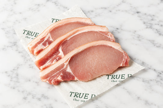 three raw pork chops with rind and visible fat layers arranged on a printed paper sheet on a dark wooden table, with a knife, red chili peppers, and string nearby.