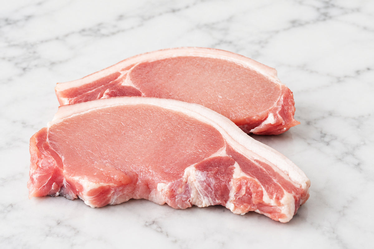 Two raw pork chops with rind and visible fat marbling, placed on a dark wooden cutting board with a knife, red chili peppers, and a small bowl nearby.