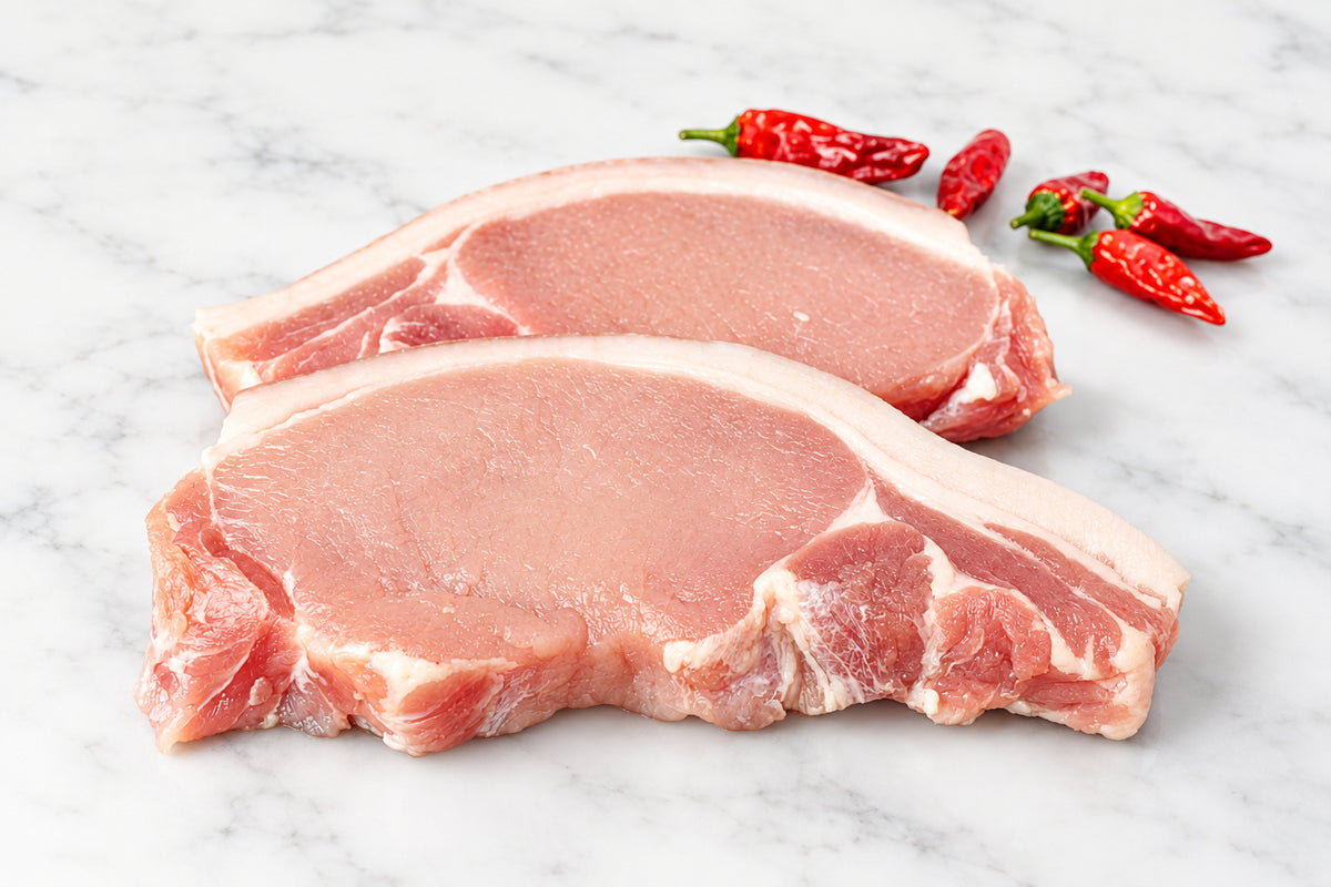 Two raw pork chops with rind and visible fat marbling resting on a dark wooden cutting board, with red chili peppers and a floral bowl in the background.