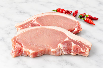 Two raw pork chops with rind and visible fat marbling resting on a dark wooden cutting board, with red chili peppers and a floral bowl in the background.