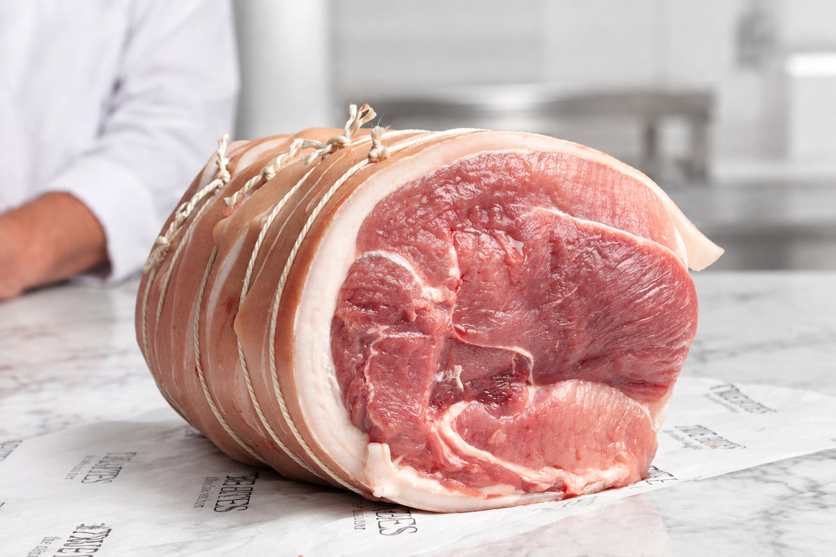 A large boneless pork shoulder tied with string, resting on branded butcher paper reading 'TRUE BITES the online butcher' on a dark countertop. A person in a white coat is partially visible in the blurred background.