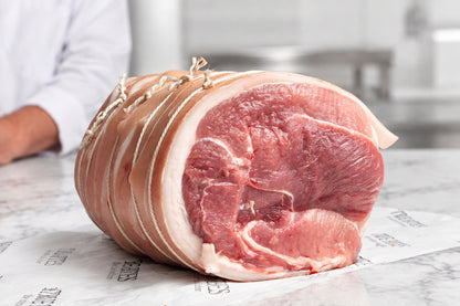 A large boneless pork shoulder tied with string, resting on branded butcher paper reading 'TRUE BITES the online butcher' on a dark countertop. A person in a white coat is partially visible in the blurred background.