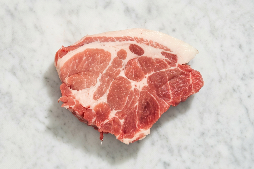 A raw cut of pork shoulder steak with visible marbling and a layer of white fat, placed on a light gray marble surface.
