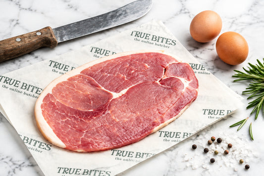 A raw cut of gammon steakresting on branded True Bites paper on a wooden table, with a knife, two brown eggs, and peppercorns nearby.
