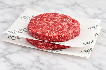 Two raw beef burger patties stacked on branded paper, placed on a wooden cutting board with coarse salt and peppercorns, with onions blurred in the background.