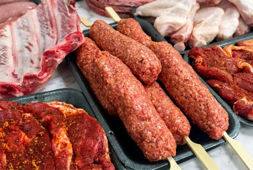 Raw meat ingredients displayed in trays, including lamb kofta kebabs on sticks, raw ribs, raw chicken pieces, and seasoned meat.