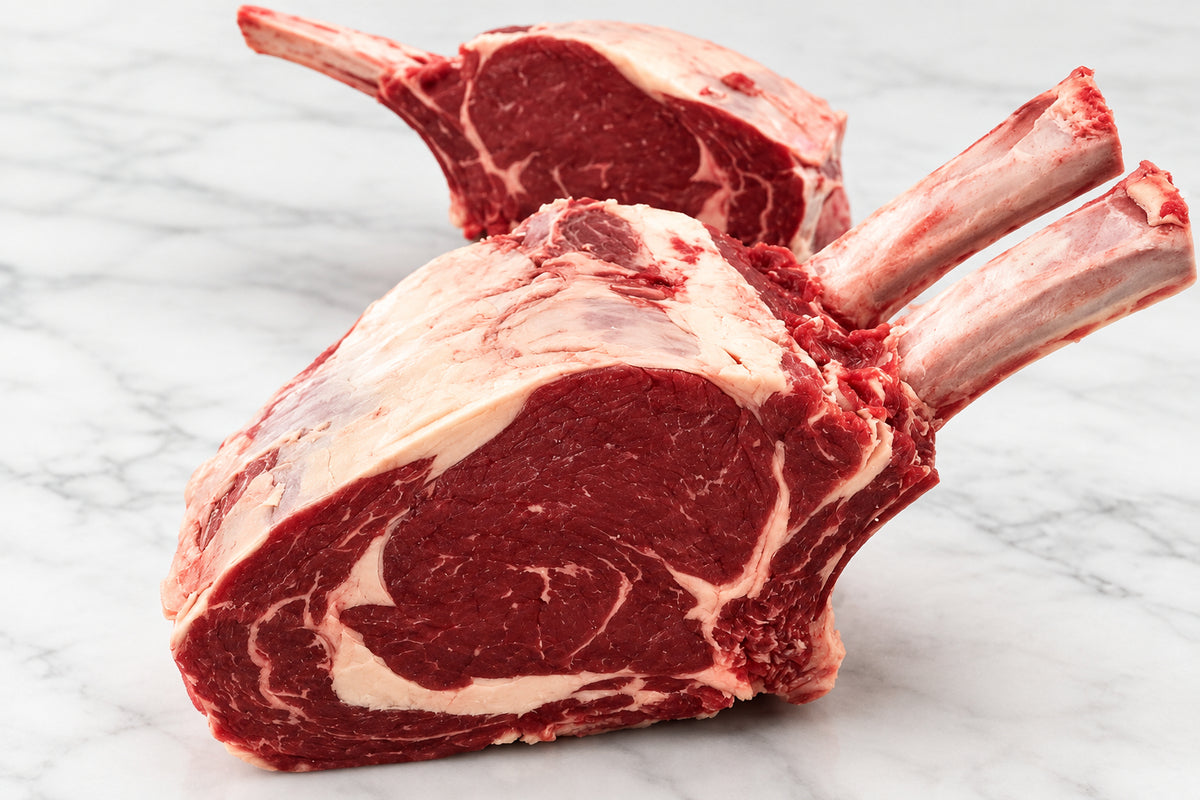 Two raw bone-in French trimmed rib of beef roasting joints with visible marbling, resting on a wooden cutting board on a stainless steel countertop.