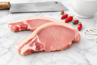 Two raw pork chops with rind and visible fat marbling on a dark wooden cutting board, with a cleaver, red chili peppers, and a ceramic bowl in the background.