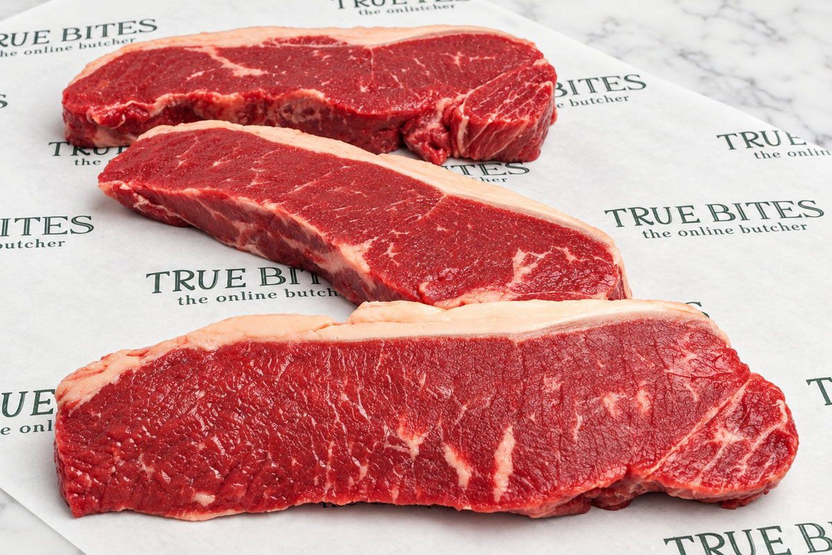 Three raw beef sirloin steaks with visible marbling, arranged diagonally on white butcher paper printed with the brand name 'True Bites Butcher'.