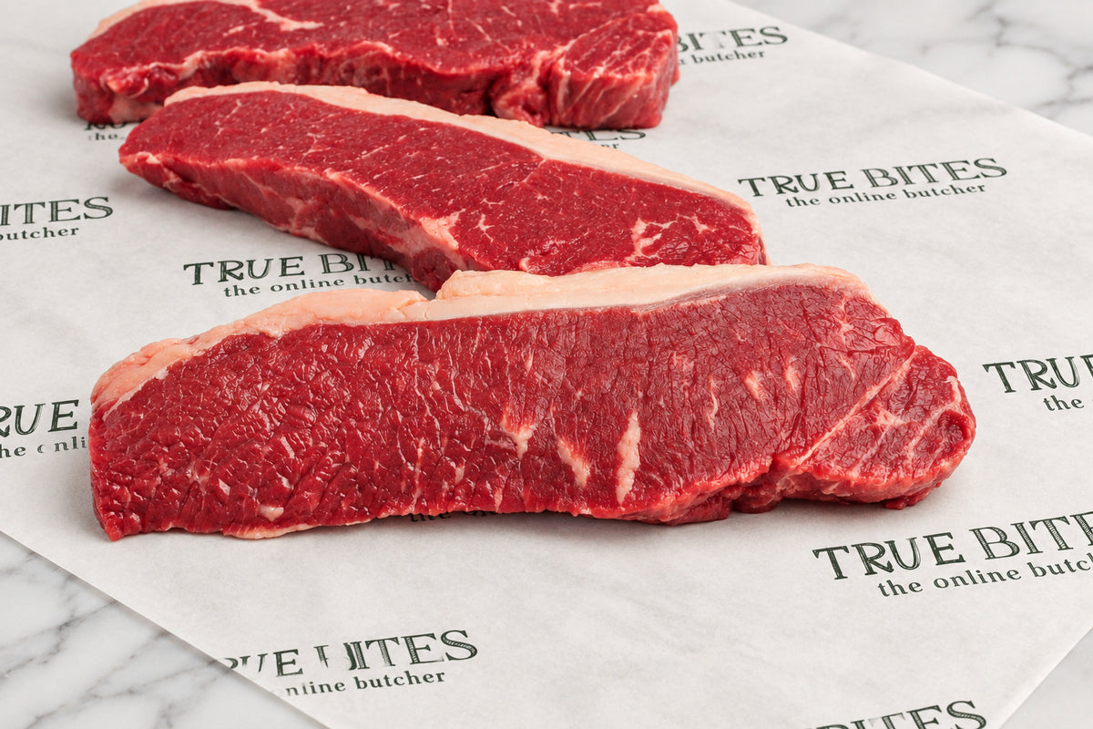Three raw beef sirloin steaks with visible fat marbling, arranged on branded butcher paper over a dark wooden surface. The paper shows the repeated brand name TRUE BITES the online butcher.