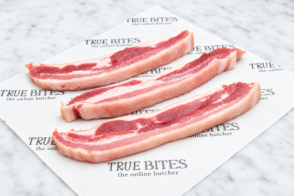 Three strips of raw sliced pork belly with rind on a 'True Bites' branded paper.