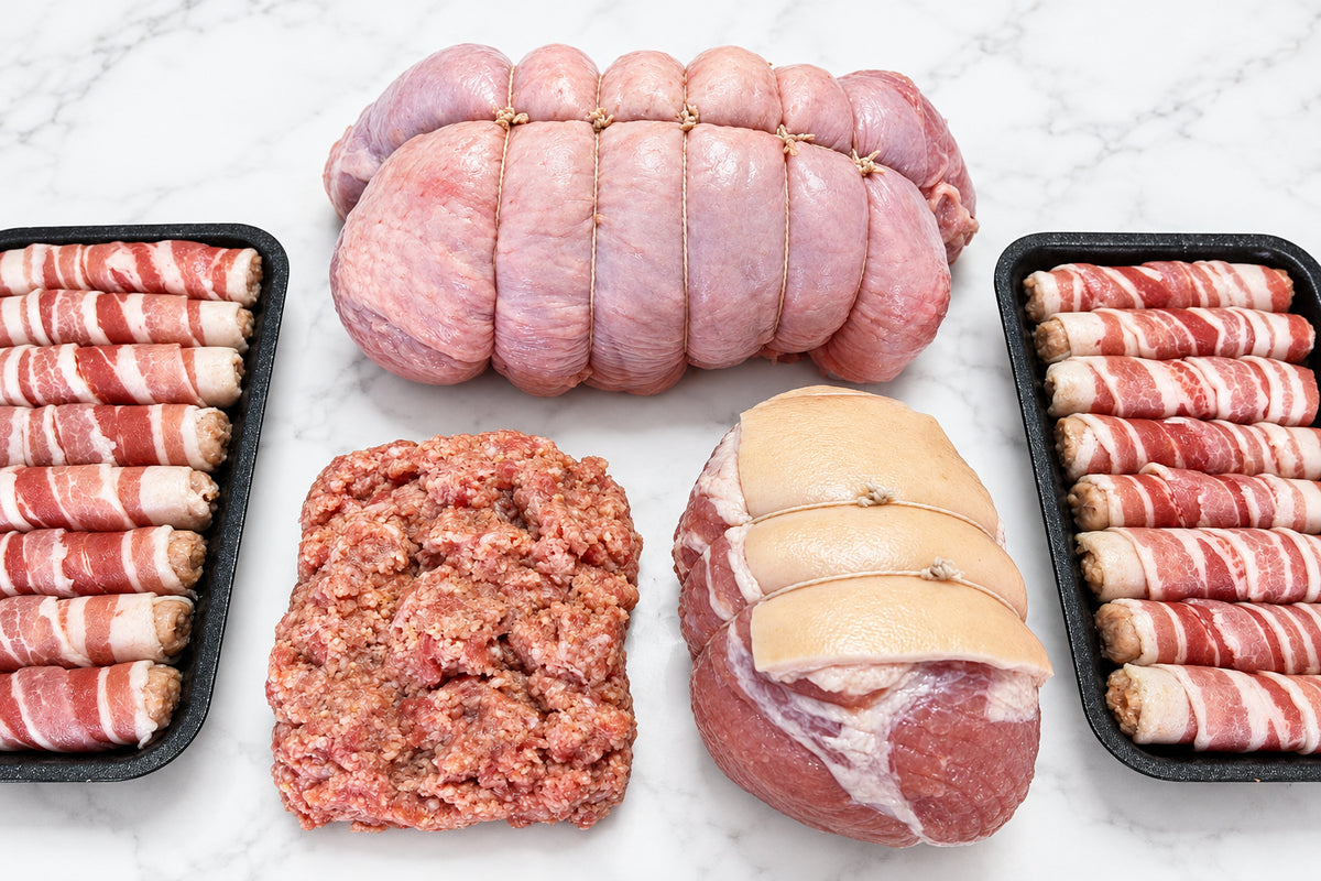 a close up of raw Christmas meats on a white surface including a strung boneless turkey breast, 2 trays of eight pigs in blankets, a gammon joint and 450g of sausage meat.