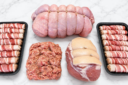 a close up of raw Christmas meats on a white surface including a strung boneless turkey breast, 2 trays of eight pigs in blankets, a gammon joint and 450g of sausage meat.