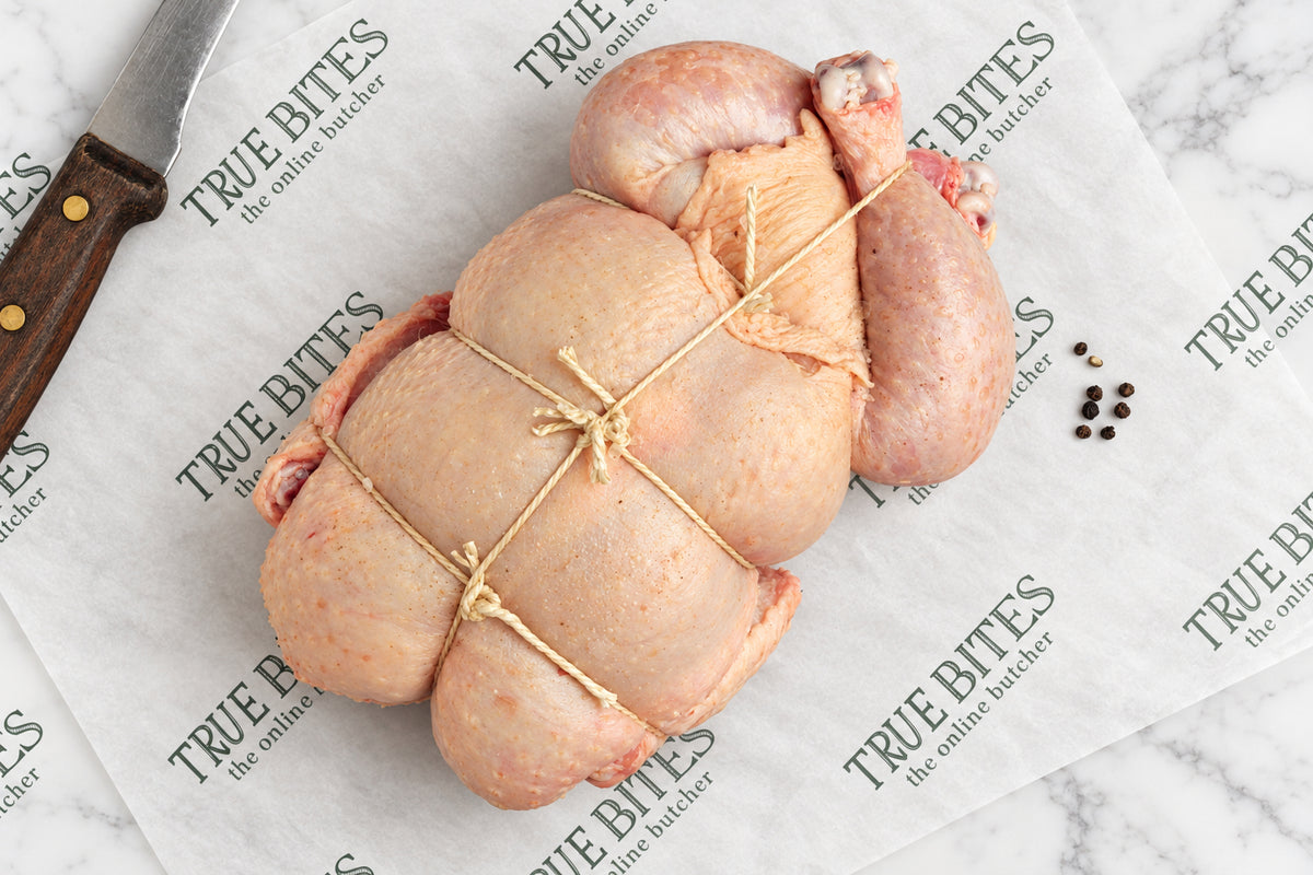 A raw, uncooked spatchcock whole chicken tied with string, placed on branded parchment paper that reads 'TRUE BITES the online butcher' with a knife and peppercorns nearby.