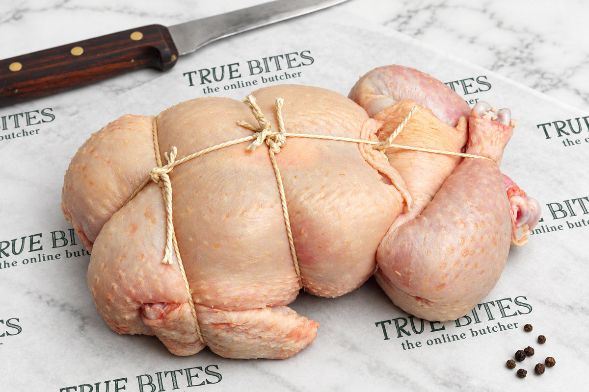 A raw, tied whole chicken placed on branded butcher paper reading 'TRUE BITES the online butcher' with a knife at the top and peppercorns at the bottom right.