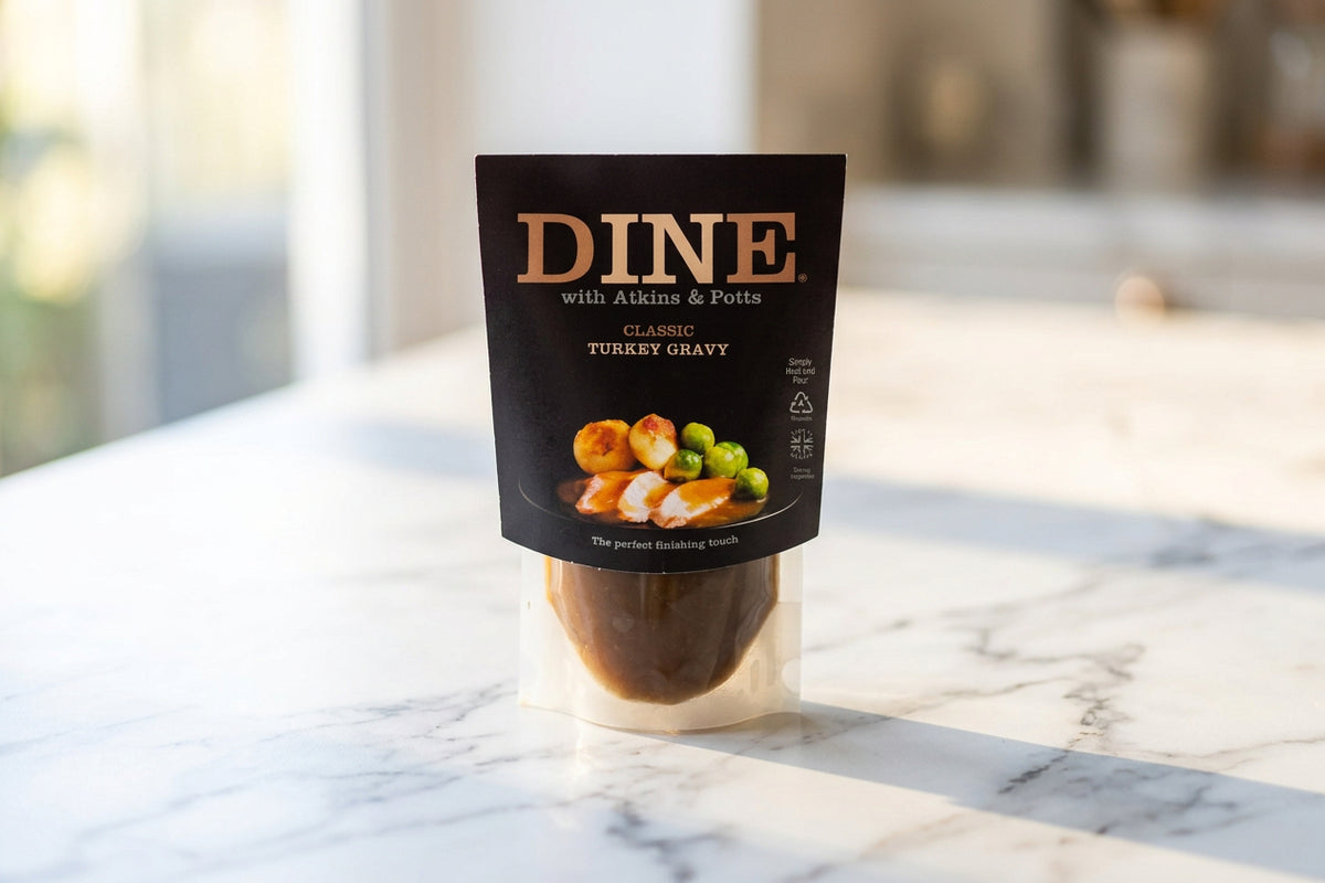 A package of DINE with Atkins & Potts Classic Turkey Gravy standing on a white desk in an office-like setting. The front label is dark brown with orange text and a food photo showing sliced turkey, carrots, and green peas.
