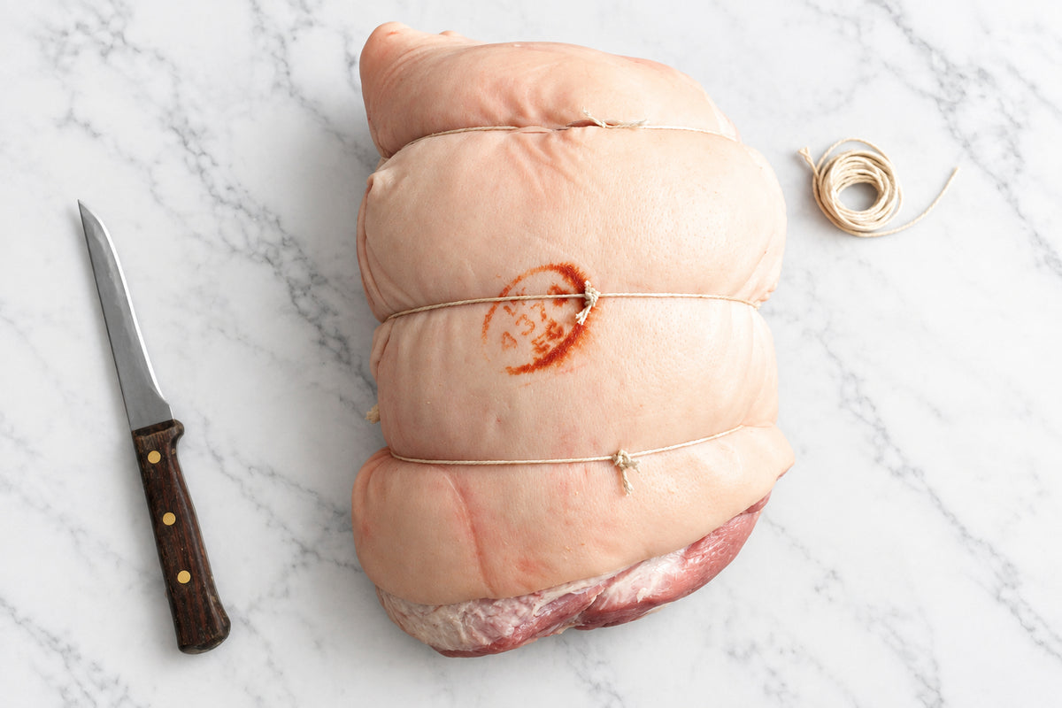 A tied raw whole boneless pork leg on a dark wooden table, with a carving knife on the left and a coil of string on the right.