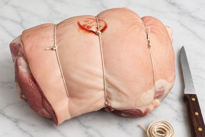 A tied raw whole pork leg roast on a dark wooden surface, with a knife and string visible beside it.