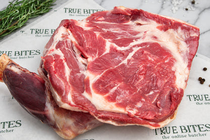 A raw whole lamb shoulder with white fat, photographed close up on printed butcher paper with herbs and peppercorns nearby.