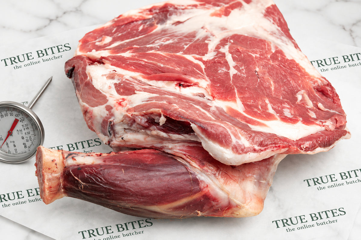 Raw whole lamb shoulder displayed on branded paper packaging with a small metal thermometer visible on the left.