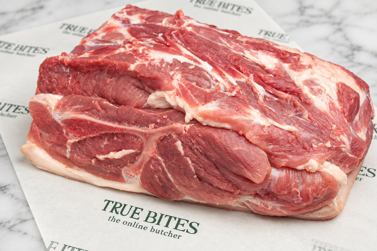 A raw whole pork butt with fat layers, placed on branded butcher paper that reads TRUE BITES the online butcher, on a dark wooden surface.