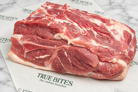 A raw whole pork butt with fat layers, placed on branded butcher paper that reads TRUE BITES the online butcher, on a dark wooden surface.