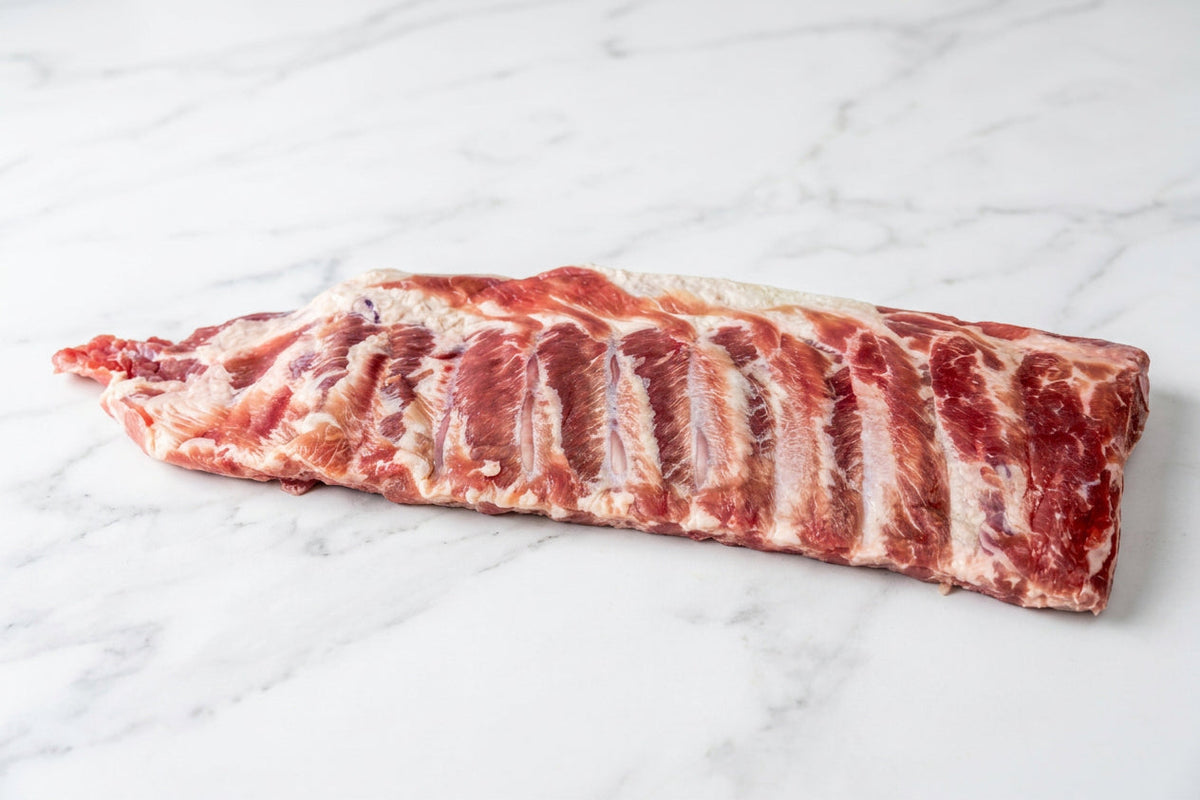 A raw whole rack of cured pork bacon ribs with visible fat marbling, placed on a white surface.