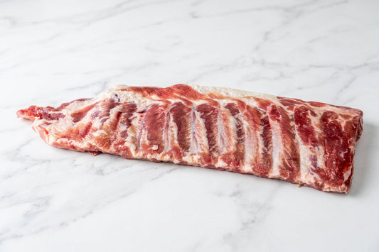 A raw whole rack of cured pork bacon ribs with visible fat marbling, placed on a white surface.