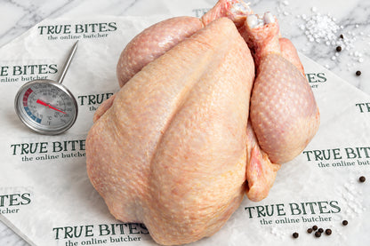 A raw whole chicken placed on branded butcher paper reading 'TRUE  BITES the online butcher' with a meat thermometer visible in the background on a dark wooden surface.