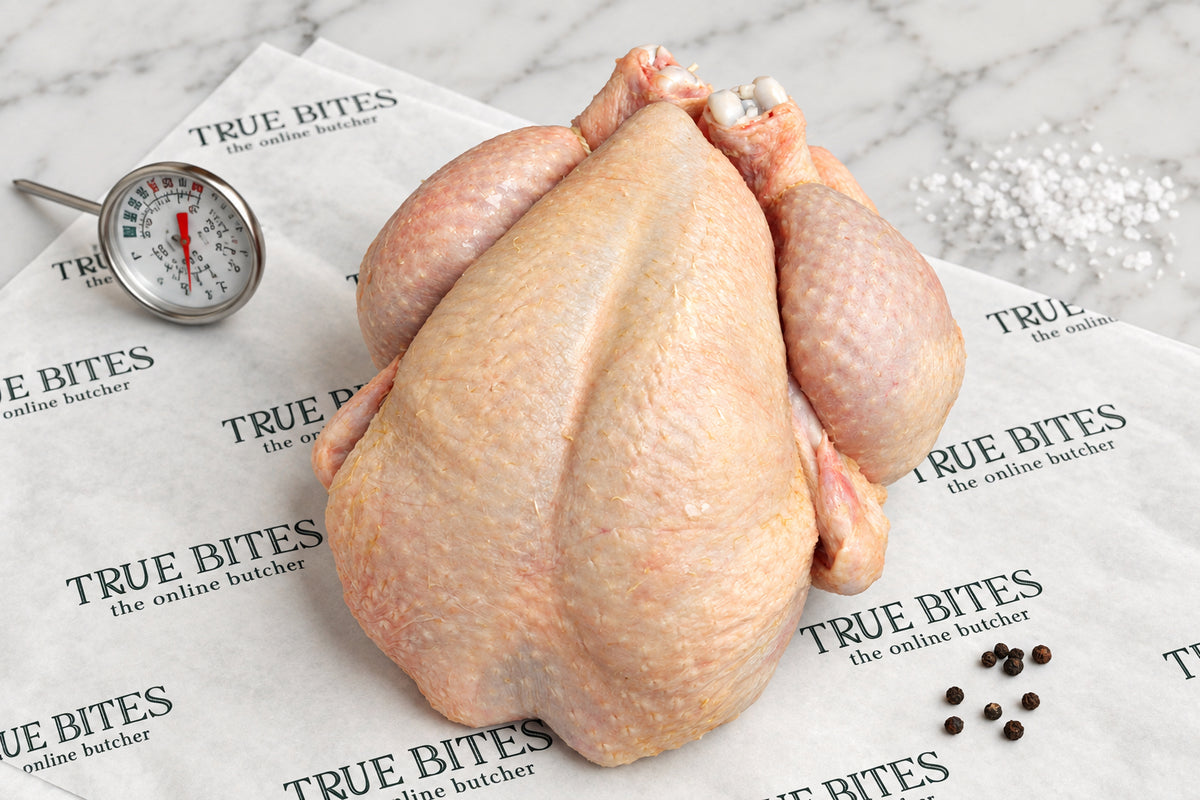 A raw whole chicken positioned upright on branded butcher paper reading 'TRUE BITES the online butcher', with a meat thermometer and peppercorns nearby on a dark wooden surface. The paper repeatedly shows the brand name TRUE BITES.