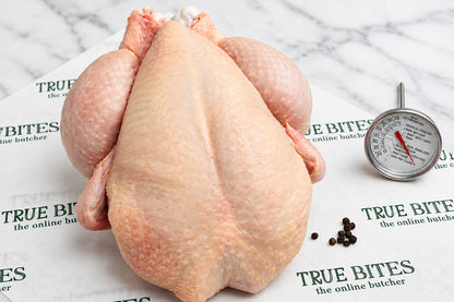 A whole raw chicken placed on branded butcher paper reading 'TRUE BITES the online butcher' with a meat thermometer and peppercorns nearby on a dark wooden surface.