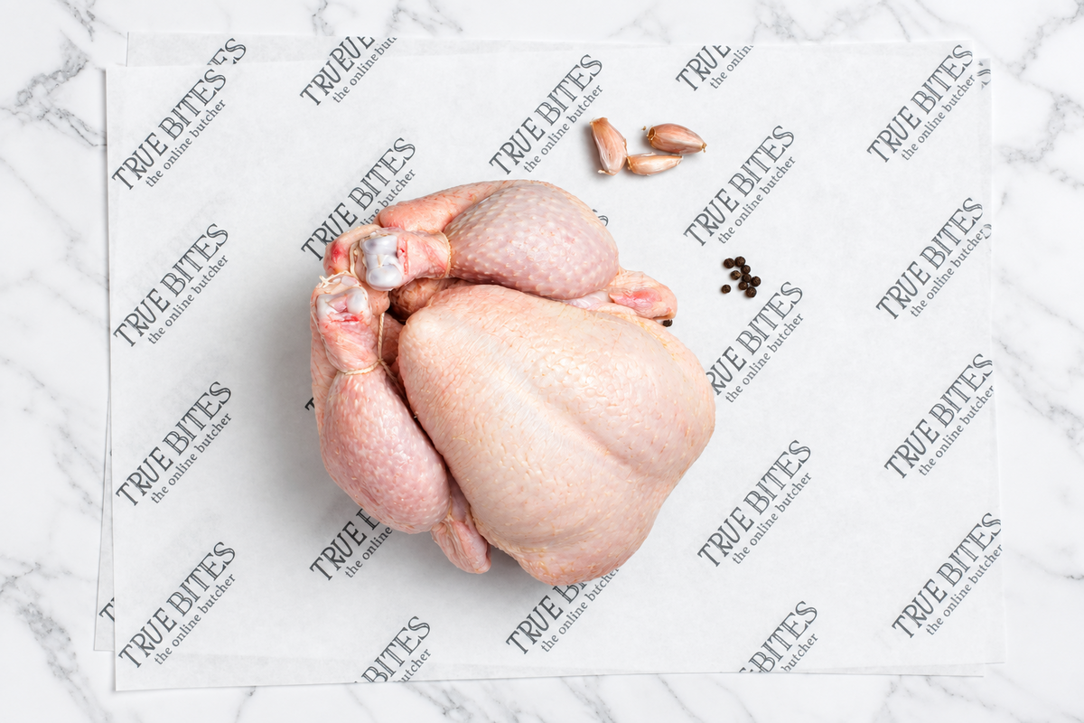 A raw whole chicken laid on branded True Bites paper, with a few garlic cloves and black peppercorns nearby on a light marble surface.