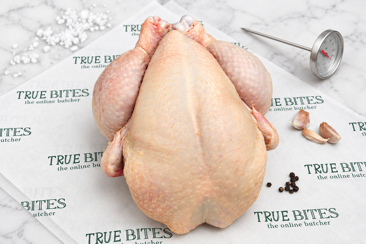 A raw whole chicken laid on branded butcher paper reading 'TRUE BITES the online butcher' with a meat thermometer, garlic cloves, and peppercorns on a dark wooden surface.