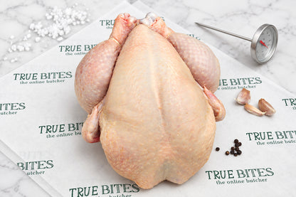 A raw whole chicken laid on branded butcher paper reading 'TRUE BITES the online butcher' with a meat thermometer, garlic cloves, and peppercorns on a dark wooden surface.
