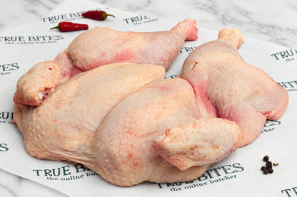 A whole raw spatchcock chicken with skin on, laid on branded butcher paper with red chili peppers and black peppercorns nearby, on a dark wooden surface.