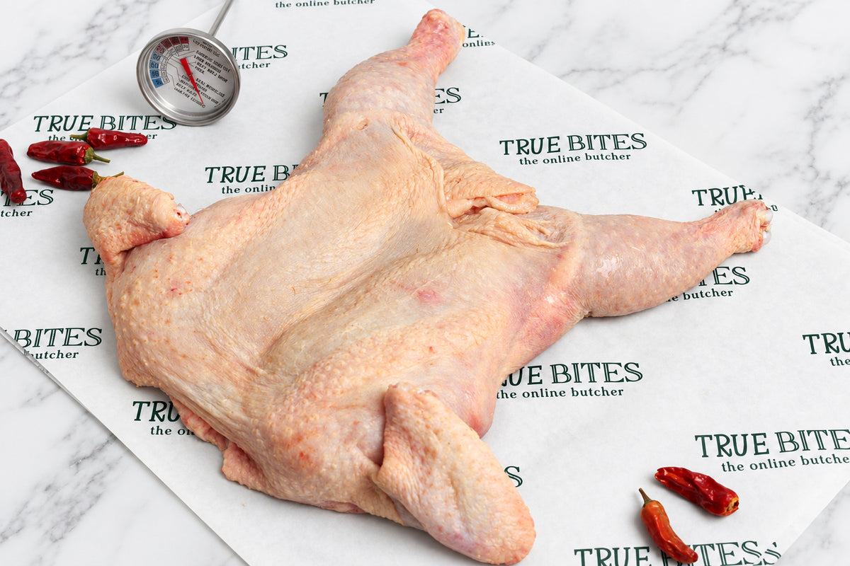 A raw, whole spatchcock skin-on chicken leg laid flat on branded butcher paper reading 'TRUE BITES the online butcher', with a metal thermometer and dried red chilies nearby on a dark wooden surface.