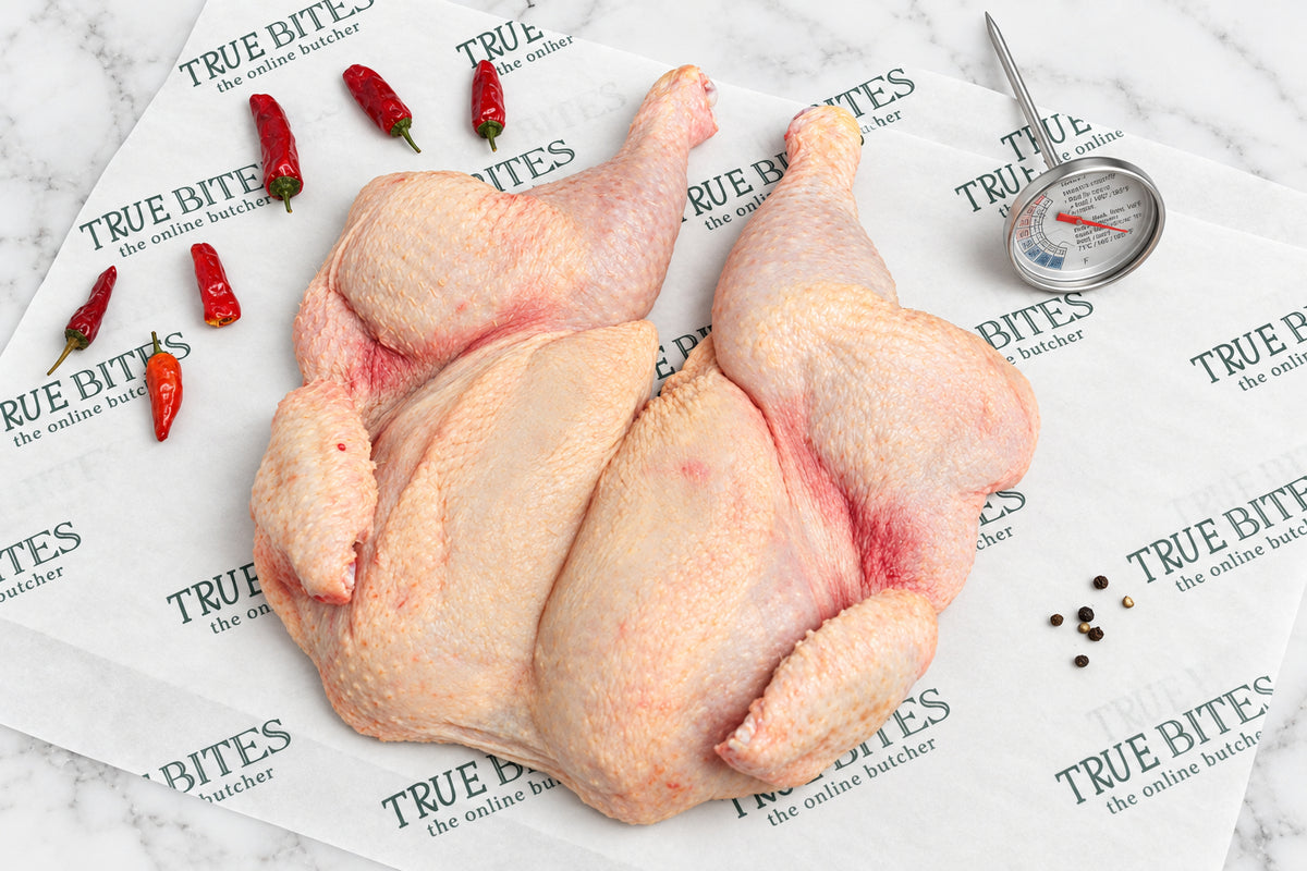 A whole raw spatchcock chicken laid on branded butcher paper reading 'TRUE BITES the online butcher' with red chili peppers, peppercorns, and a meat thermometer nearby on a dark wooden surface.