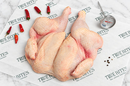 A whole raw spatchcock chicken laid on branded butcher paper reading 'TRUE BITES the online butcher' with red chili peppers, peppercorns, and a meat thermometer nearby on a dark wooden surface.