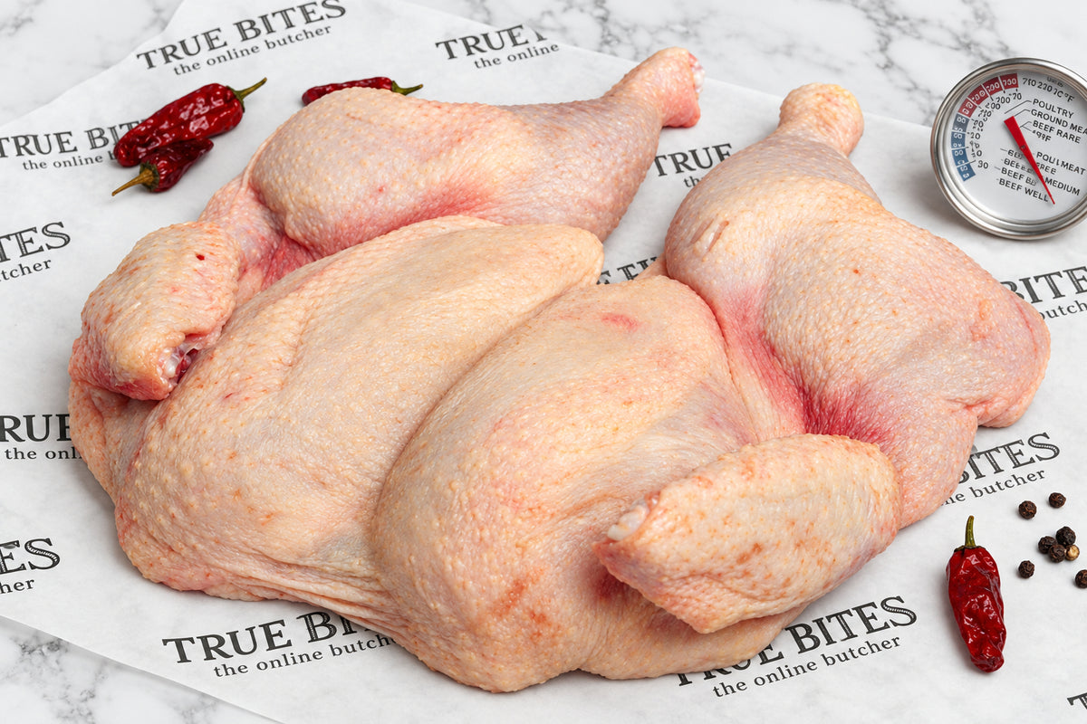 Raw whole spatchcock chicken pieces arranged on branded butcher paper reading 'TRUE BITES the online butcher' with red chili peppers and a thermometer visible in the scene.