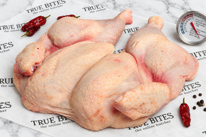 Raw whole spatchcock chicken pieces arranged on branded butcher paper reading 'TRUE BITES the online butcher' with red chili peppers and a thermometer visible in the scene.