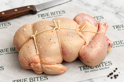A raw, tied whole chicken placed on branded butcher paper reading 'TRUE BITES the online butcher' with a knife and peppercorns nearby, photographed in a real kitchen/butcher setting rather than a clean studio packshot.