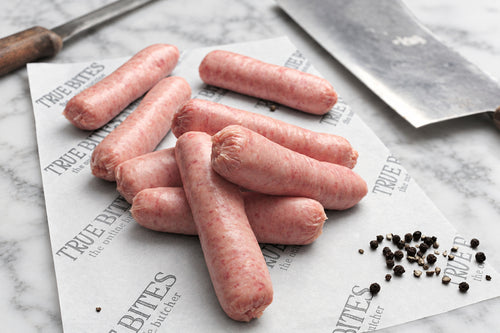 8 raw pork sausages on branded 'TRUE BITES the online butcher' butcher paper with black pepper and butchers knives on table